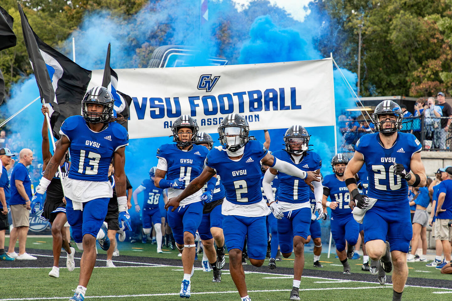 A group of football players in blue uniforms run onto the field with smoke and a banner reading "GVSU FOOTBALL" behind them.